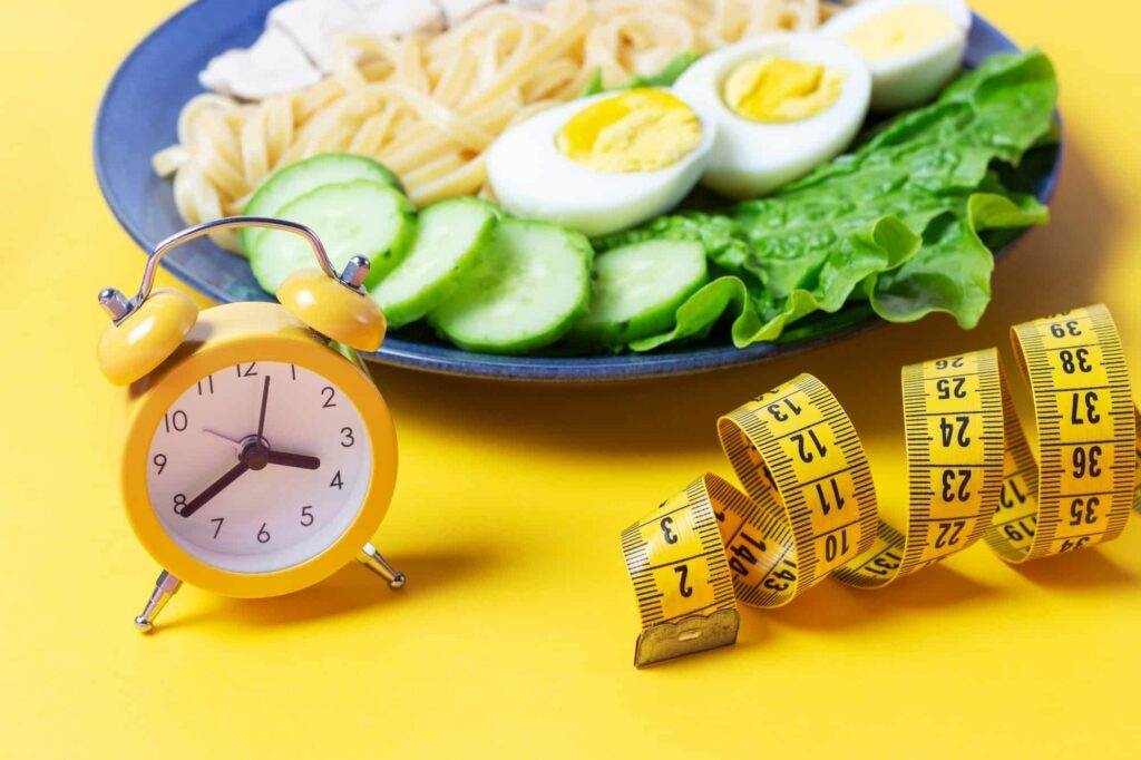 Plate with food and alarm clock on yellow background, interval fasting concept