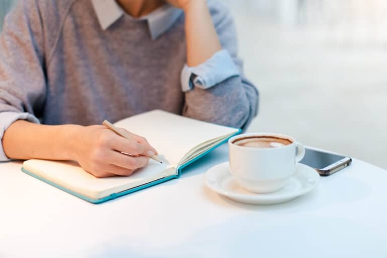 Young woman is writing in notebook in cozy cafe with cup of coffee. Freelancer is working in comfortable coworking modern place. Girl is planning and dreaming. Wellbeing in workplace.