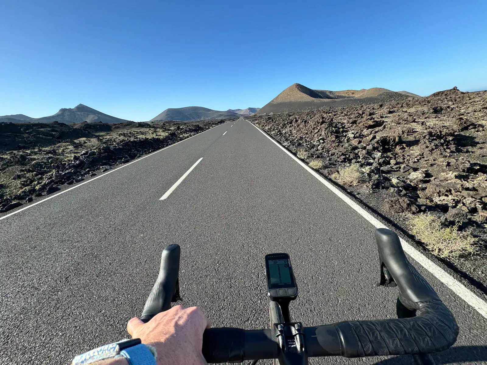 Cyclist navigates a deserted road through Lanzarote's rocky, mountainous landscape beneath a clear blue sky.