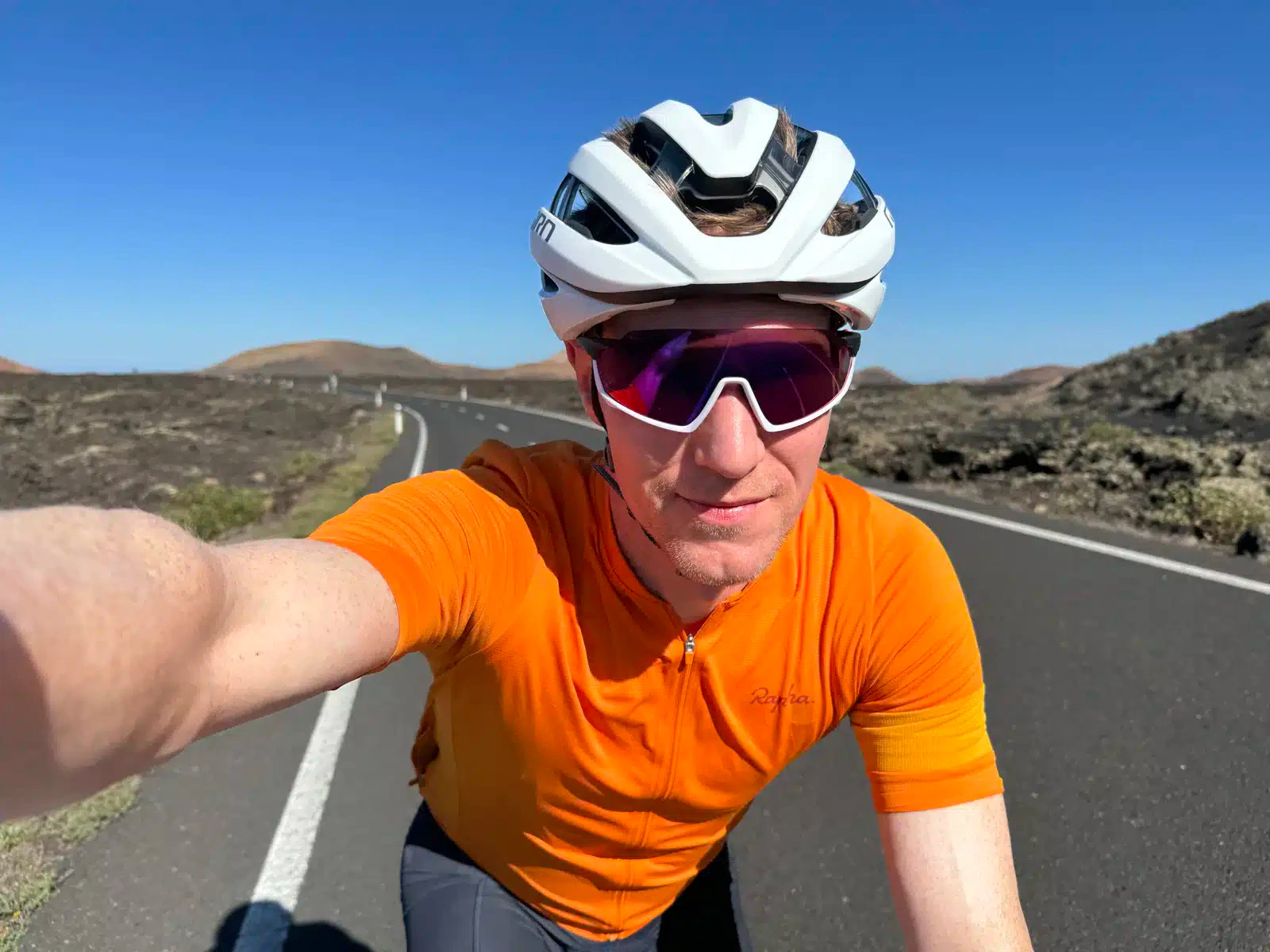 A cyclist in a helmet and sunglasses taking a selfie on a sunny road.