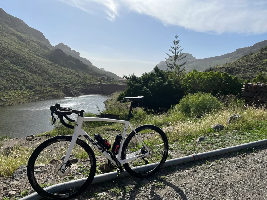 In Gran Canaria, a white road bike sits beside the roadside against a backdrop of mountain peaks and a shimmering reservoir.