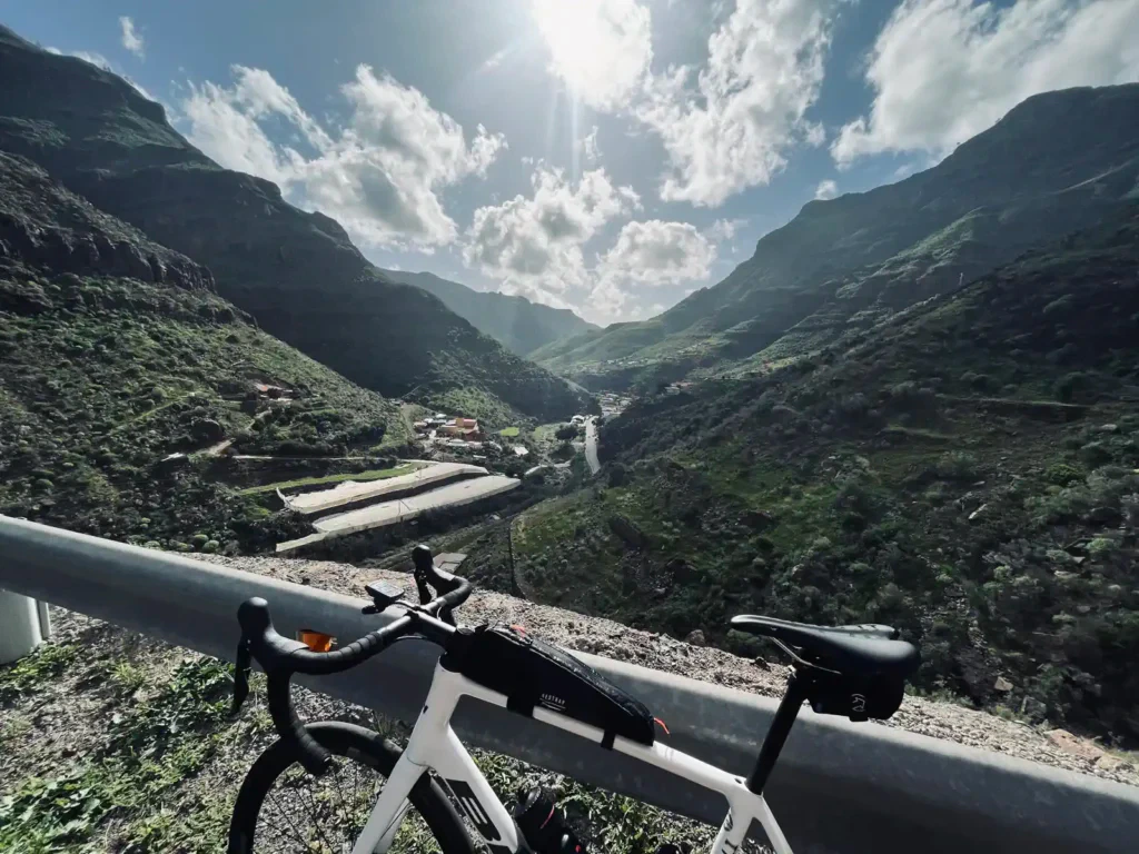 A road bike leaning on a guardrail overlooking a sunlit mountainous landscape with a small village nestled in a valley.