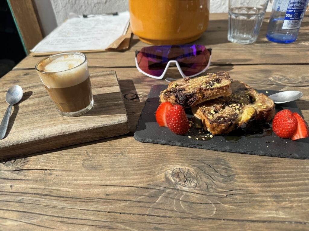 A small glass of coffee and a spoon sit on a wooden tray beside a slate platter with two pieces of toasted bread topped with seeds and accompanied by strawberries. Sunglasses and a drink pitcher are in the background.