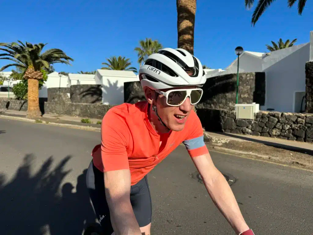 A cyclist in an orange jersey and white helmet rides down a sunny street lined with palm trees and white buildings.