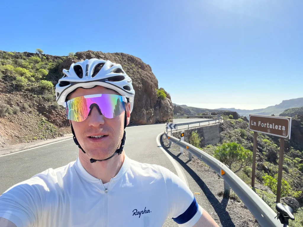 Cyclist in Gran Canaria, with helmet and reflective glasses, stops on a sunlit mountain road past a "La Fortaleza" sign.