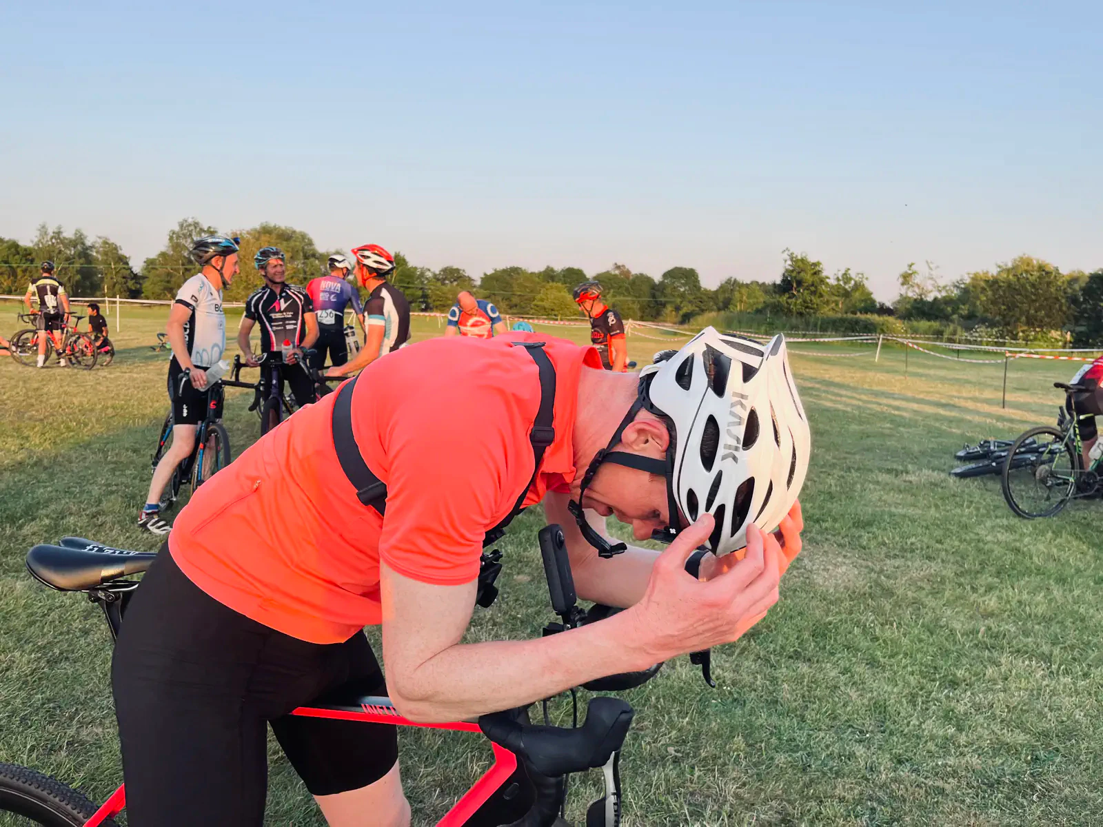 A cyclist in an orange shirt adjusts their helmet while standing next to a red bicycle. Other cyclists are gathered in the background on a grassy field.