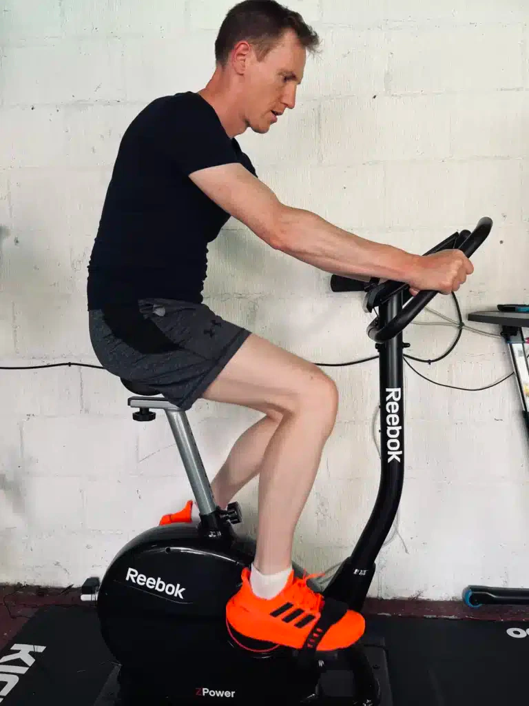 A person in a black shirt and gray shorts rides a Reebok stationary bike indoors. They are wearing bright orange athletic shoes.