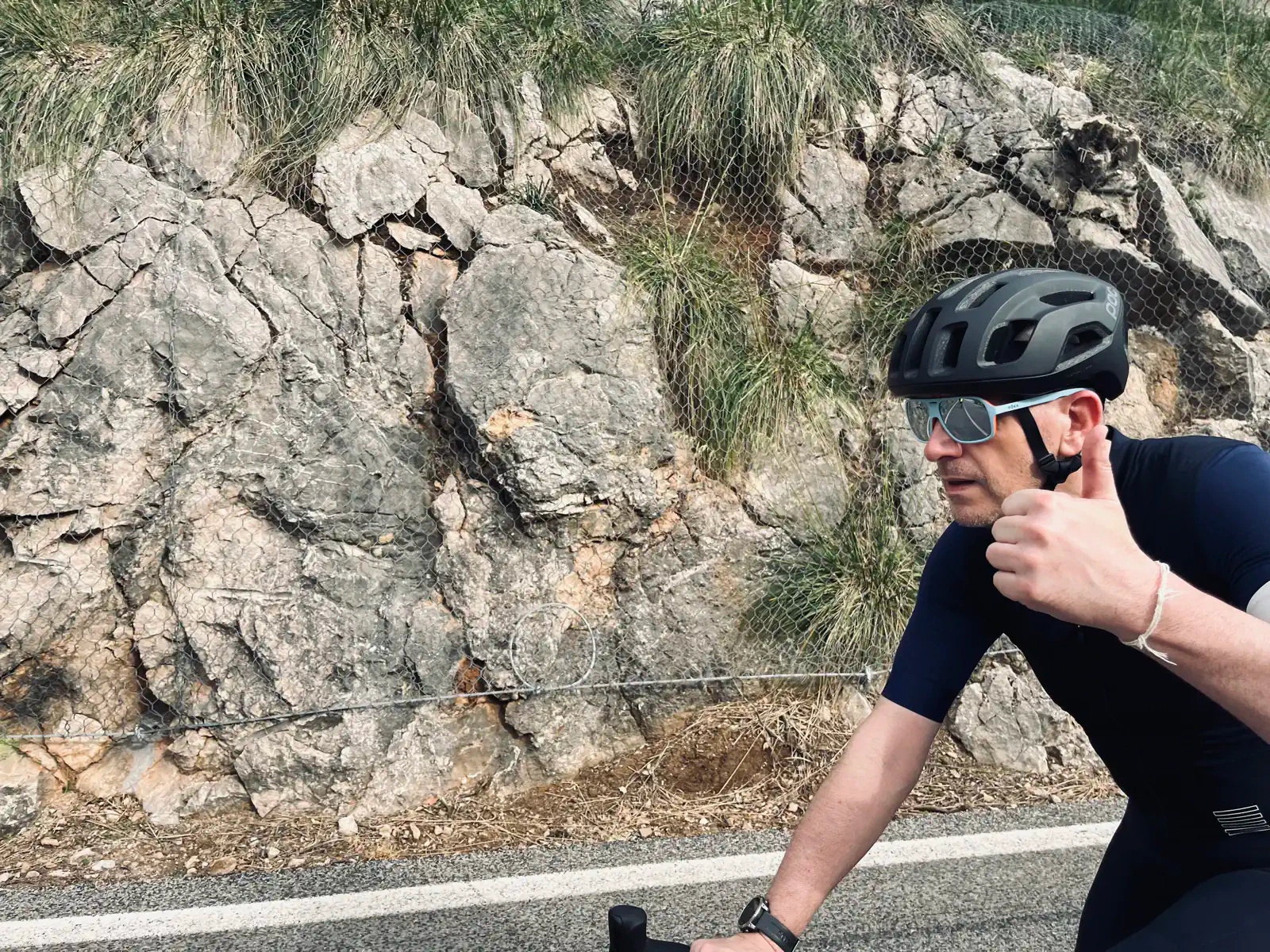 A cyclist in a black helmet and shades gives a thumbs-up while riding by a rocky hillside.