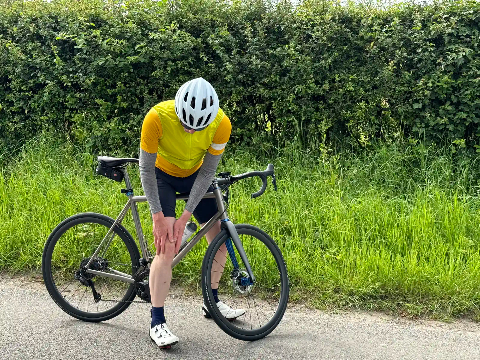 A cyclist in a yellow jersey and white helmet is bent over their bike, clutching their knee, on a road next to some thick bushes.
