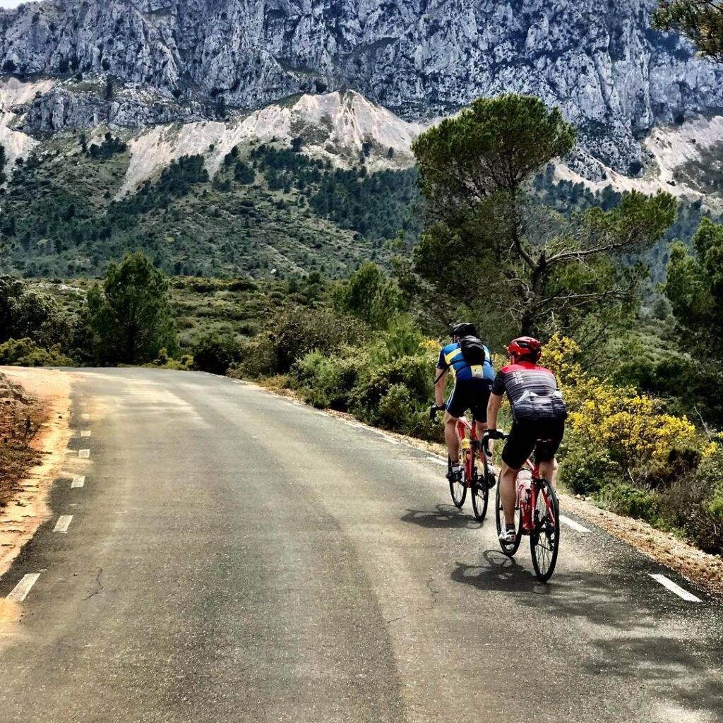 Two cyclists tackle a scenic, challenging route near Denia, surrounded by lush greenery and rocky cliffs under clear skies—a prime location for cycling holidays.