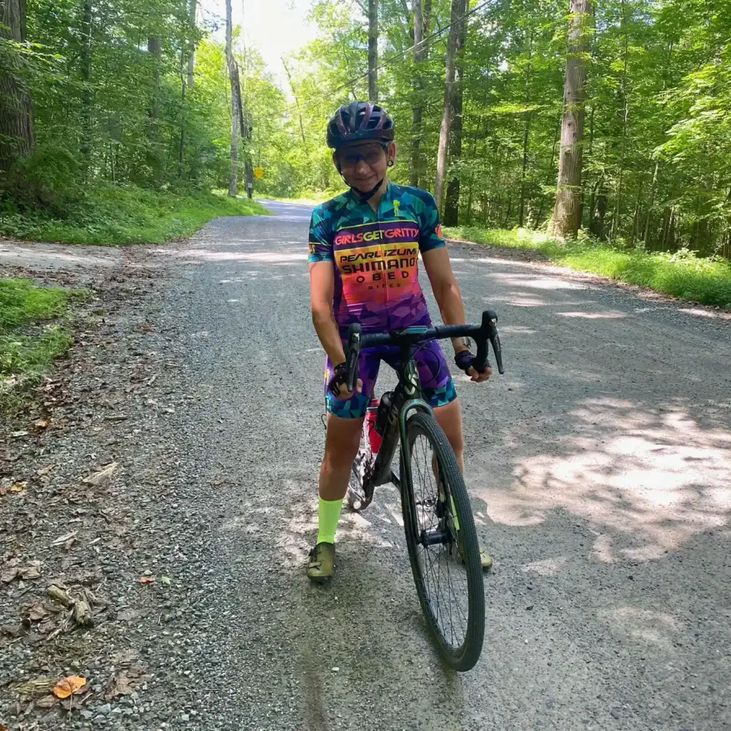Amanda cycling for pedallers Amanda in colorful attire standing with a bicycle on a shady forest road.