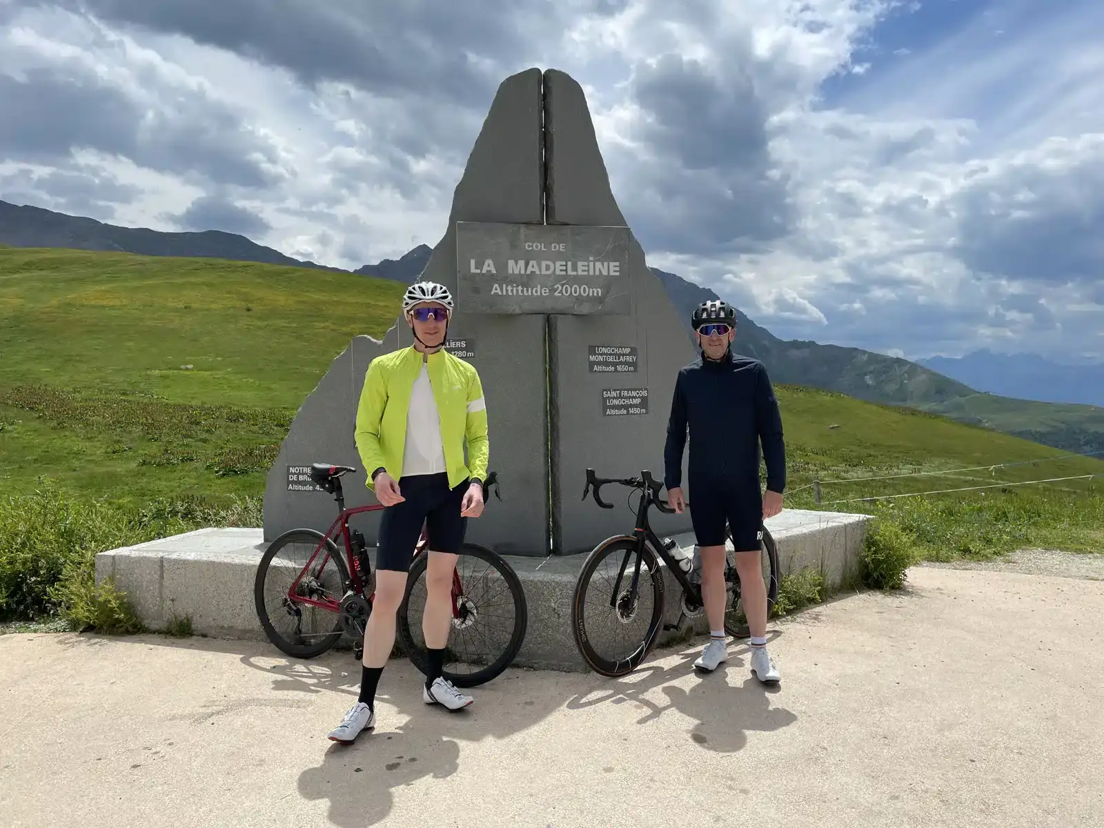 Two cyclists standing by a monument at col de la madeleine, at an altitude of 2000m, with scenic mountain views in the background. Two cyclists standing by a monument at col de la madeleine, at an altitude of 2000m, with scenic mountain views in the background.