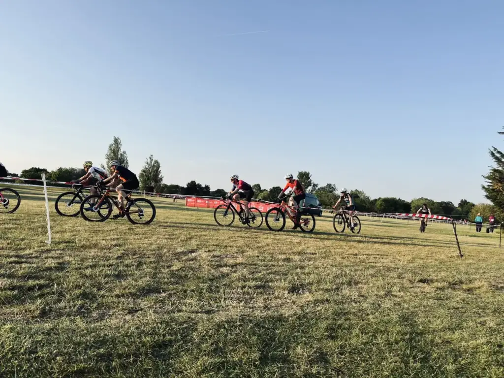 A group of cyclists are riding on a grassy field, participating in what appears to be a cycling event. The sky is clear, and the field is bordered by red and white tape. Trees are visible in the background.