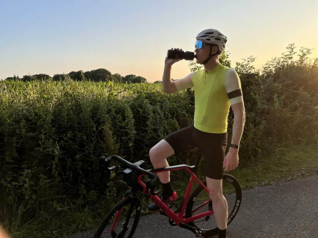 There's this cyclist in a yellow jersey and black shorts just chilling next to their red and black bike, sipping on some water. The scenery's all green and sunny.