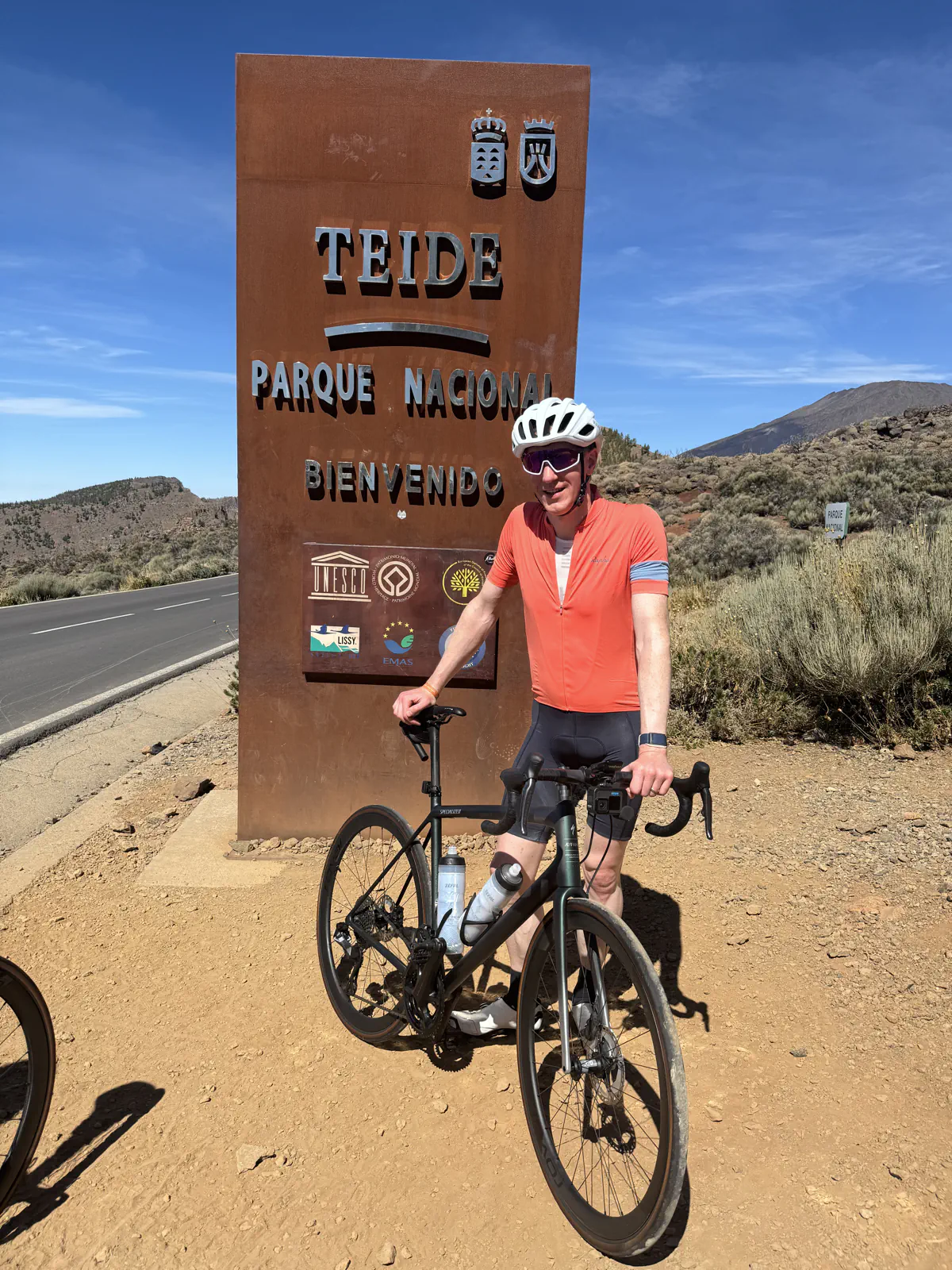 Cyclist in an orange jersey strikes a pose with their bike next to the Teide National Park sign under sunny skies.