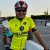 Cyclist in a bright yellow jersey and helmet, smiling with a water bottle in hand, stands confidently next to his bike by a racetrack.