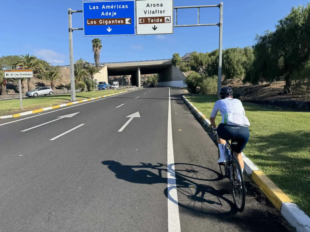 Cyclist rides under the sun past signs for Las Américas, Los Gigantes, Arona, and El Teide with a bridge looming ahead.